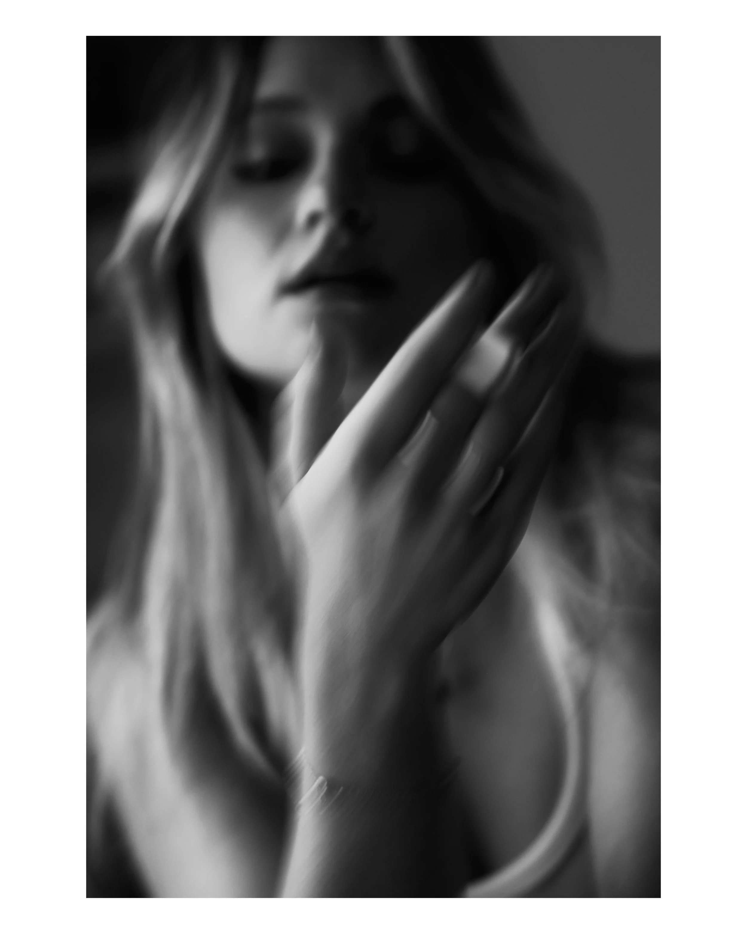 Black and white close-up of a woman with long hair, holding her face gently with her hand, looking softly at the camera.