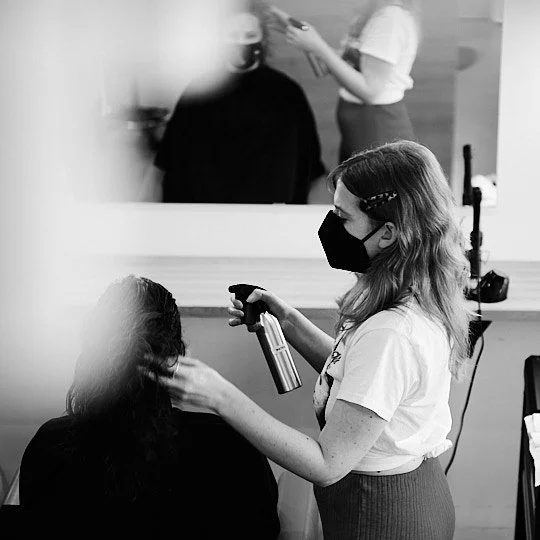A hairstylist wearing a face mask sprays hair of a client sitting in a salon chair, with a mirror reflecting other salon staff and clients.