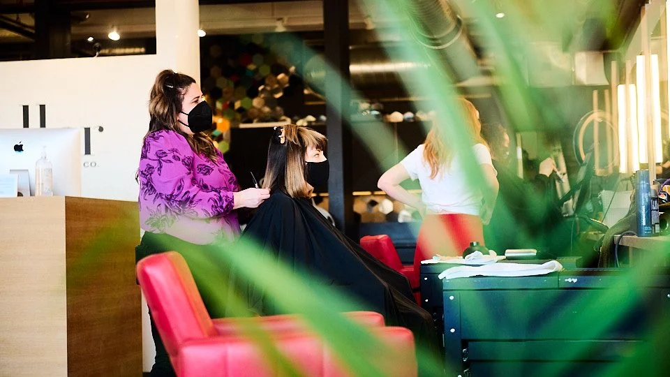 A woman with long brown hair and a black face mask is standing behind a seated woman with shoulder-length brown hair, also with a black face mask, inside a hair salon. The stylist is wearing a purple top with floral patterns, and the client is covere