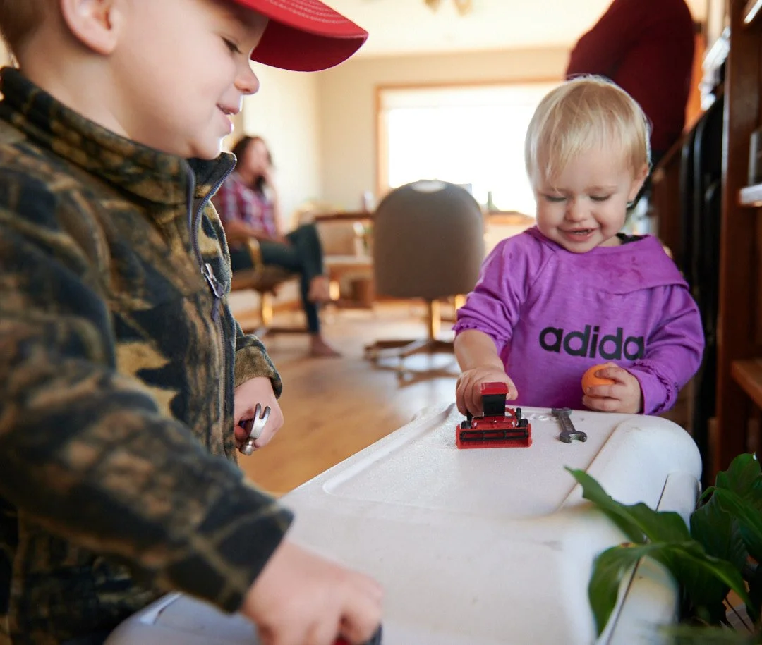 Two young children, a boy and a girl, are standing at a table with tools in their hands. The boy is wearing a camouflage jacket and a red hat, while the girl is wearing a purple Adidas sweatshirt. They are playing with a red stapler on the table, smi