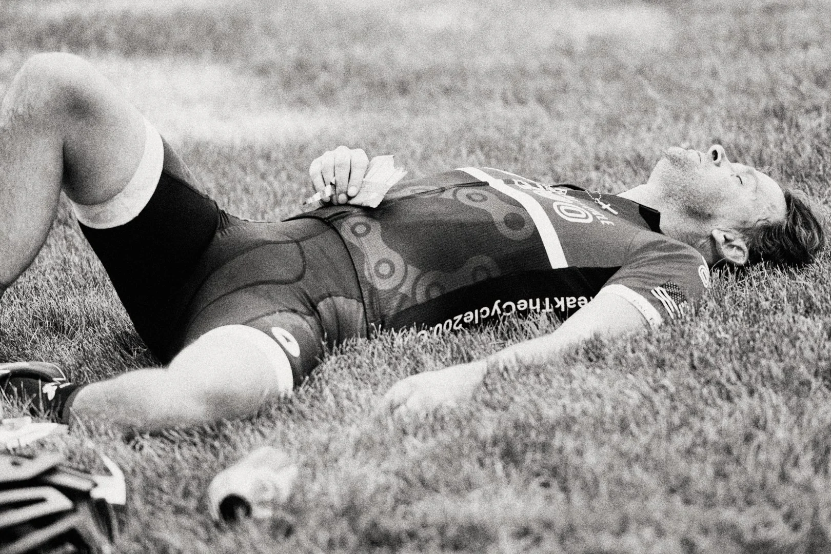 A cyclist lying on the grass after a race, holding a paper towel and looking relaxed.