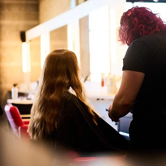 A woman with long wavy hair sitting in a salon chair, getting her hair styled by a stylist with short, curly pink hair, in a well-lit salon