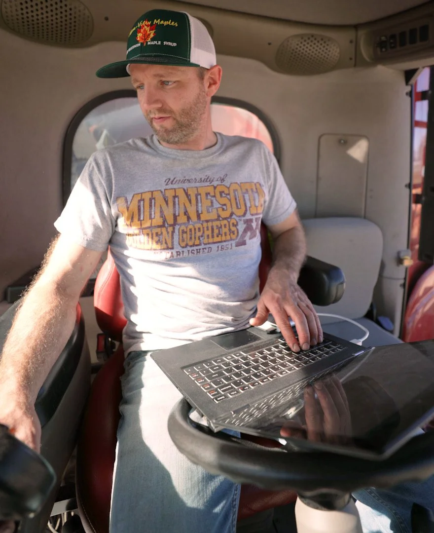A man with a beard wearing a gray T-shirt with 'University of Minnesota Golden Gophers' written on it, and a green and white trucker hat with 'Vimy Maples' and an orange maple leaf embroidered on it, sitting inside a vehicle cab working on a laptop p