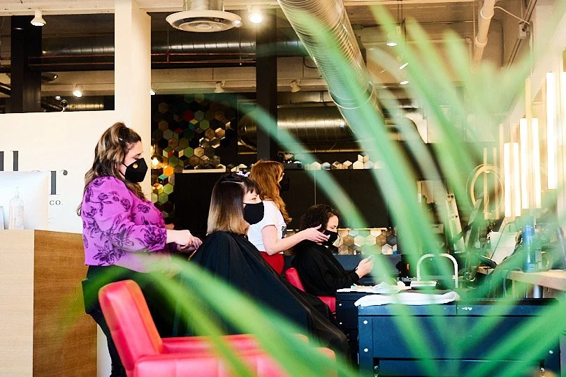 Inside a modern hair salon with four clients seated and getting hair services, all wearing face masks, with decor including hexagonal tiles on the wall and potted plants.