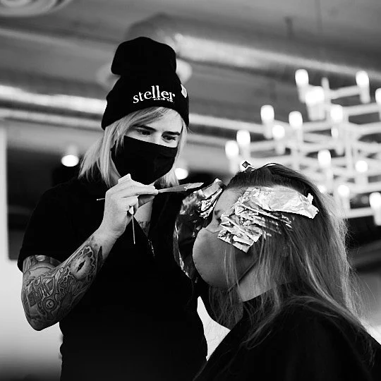 Hairdresser applying foil highlights to a client's hair in a salon, both wearing masks.