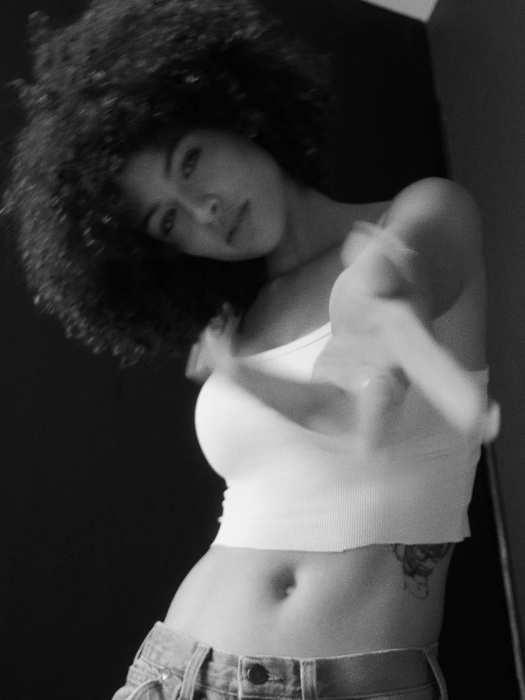 Black and white selfie of a woman with curly hair in a white tank top and jeans, taking a mirror selfie with her arm extended.