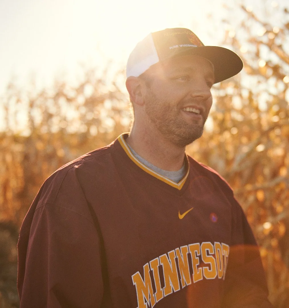 A man wearing a Minnesota sports jersey and a cap, standing outdoors during sunset with trees in the background.
