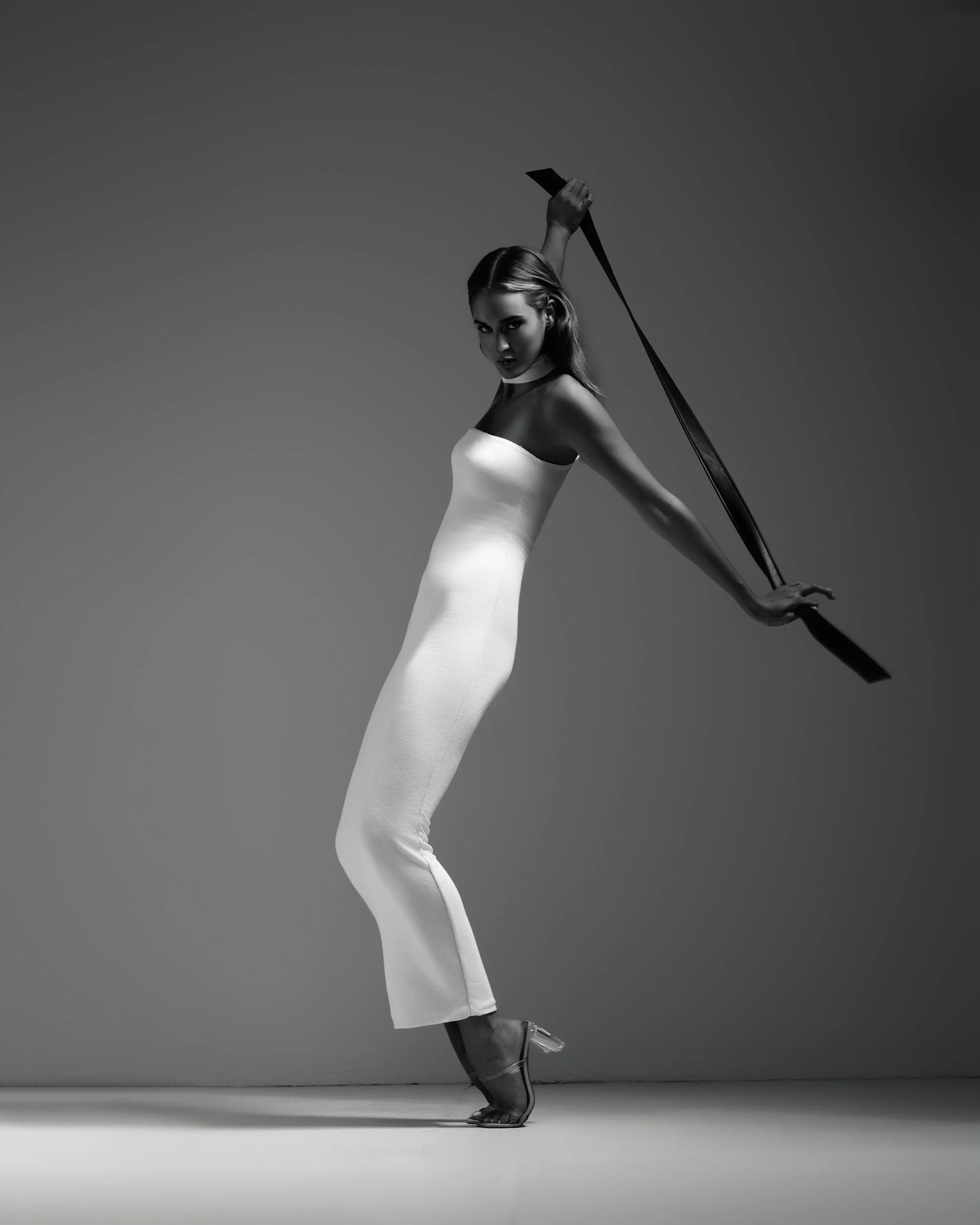 A woman in a white strapless dress and clear high heels poses against a plain background, holding a long black pole extended behind her back.