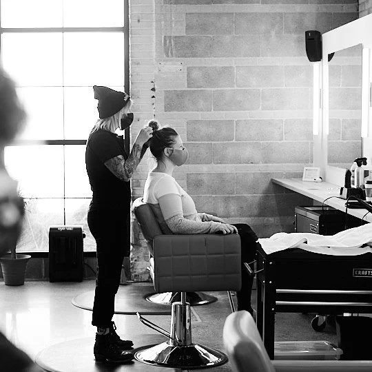 A hairstylist working on a woman's hair in a salon with a large window in the background.