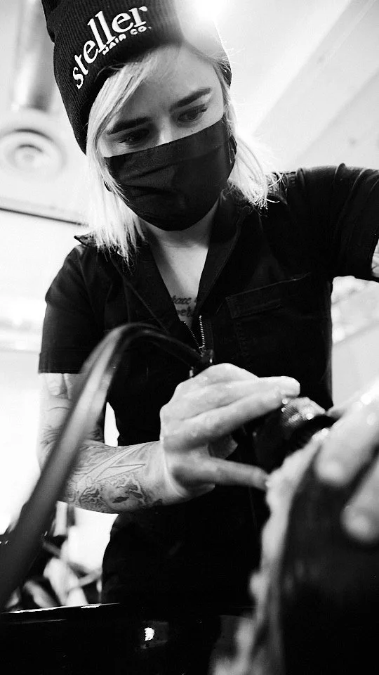 A woman with blonde hair, wearing a beanie with 'Steller' embroidered on it, a black face mask, and black clothing, taking a close-up photo with a camera. She has tattoos on her left arm.