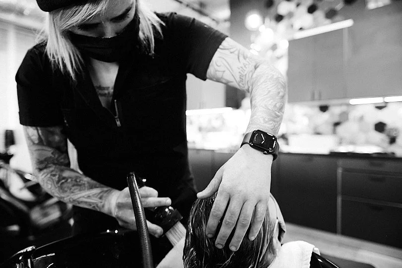 A hairstylist washing a client's hair in a salon, with tattoos on her arms and wearing a smartwatch.