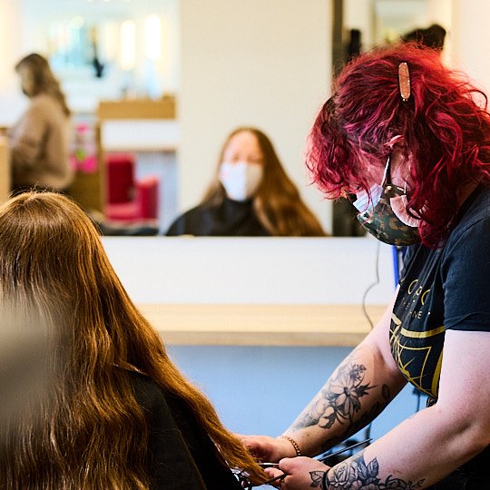 A tattooed woman with red hair and glasses wearing a face mask is giving a vaccination to a woman with brown hair in a public health setting.