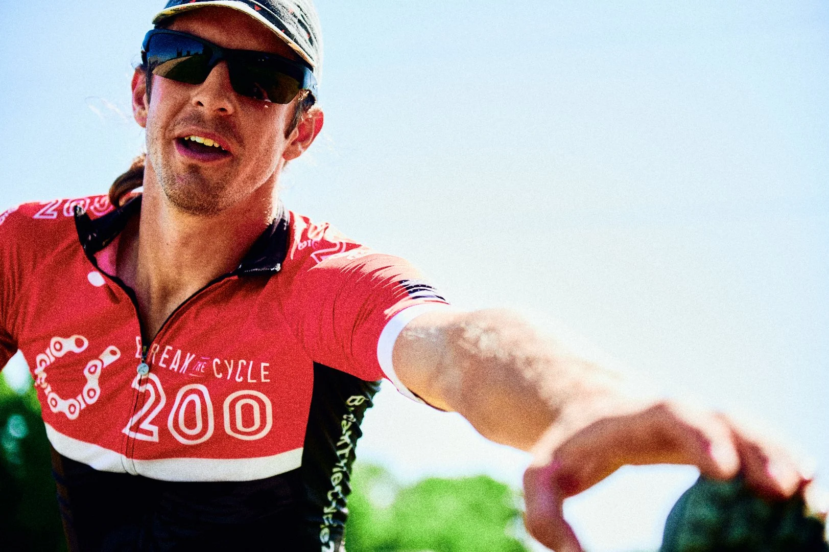 Close-up of a man with dark sunglasses and a cycling cap, wearing a red cycling jersey with white and black accents. The man is taking a selfie during a bike ride, reaching out with his arm towards the camera, with a clear blue sky in the background.