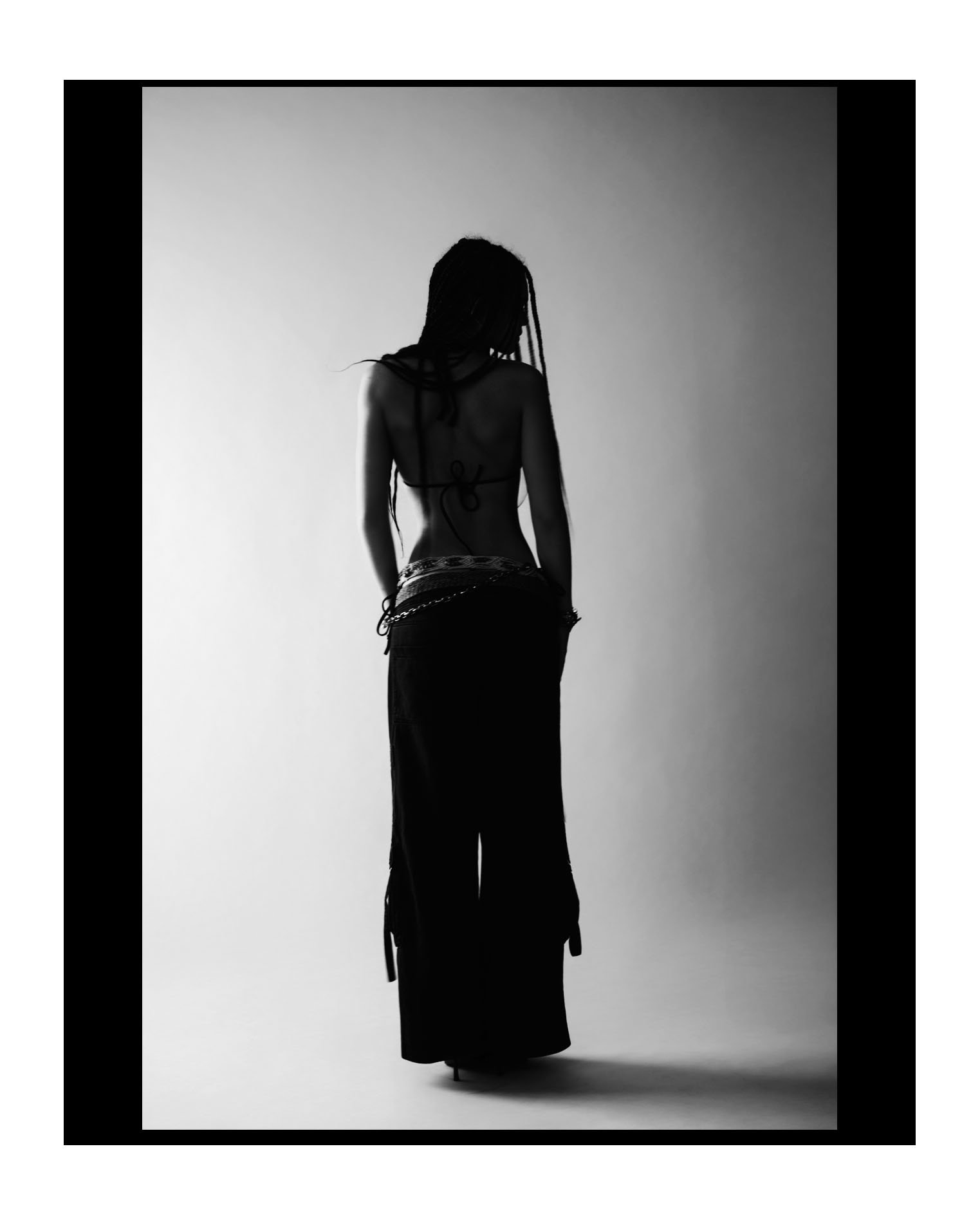 Silhouette of a woman with dreadlocks, wearing a backless top and long skirt, standing against a gradient background with a black border.