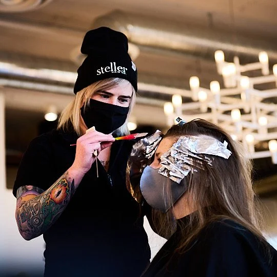 A hairstylist applying hair foil highlights to a woman with long brown hair, both wearing masks indoors.