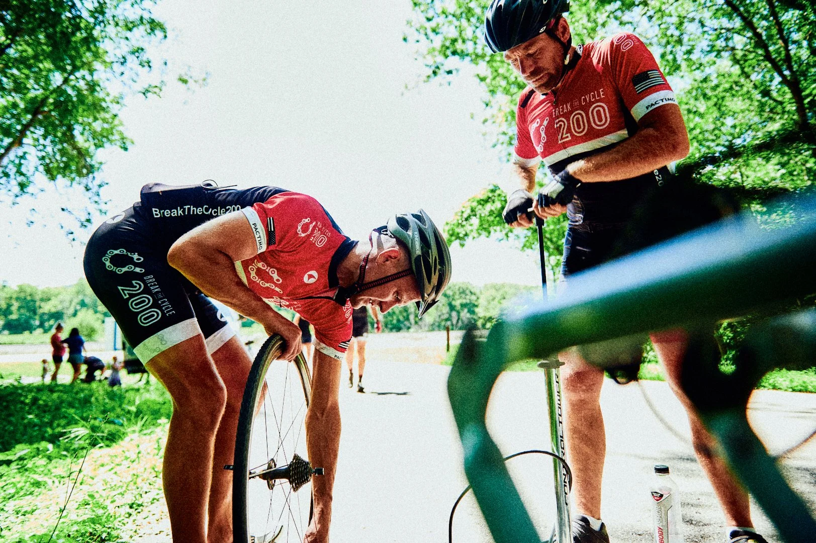 Two cyclists wearing red jerseys and helmets repairing a flat tire on the side of a sunny park trail, with trees and other cyclists in the background.