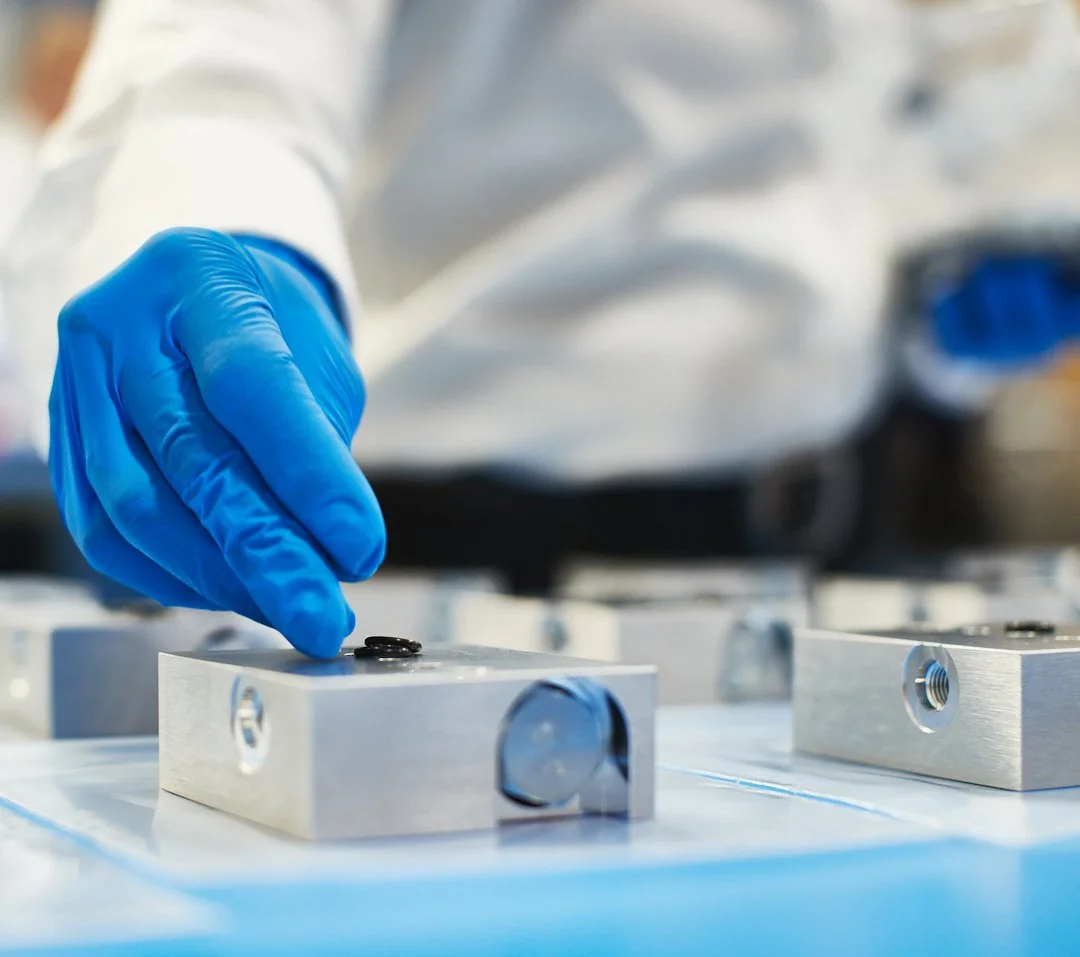 A scientist wearing blue gloves and a white coat working with metal components on a workbench.