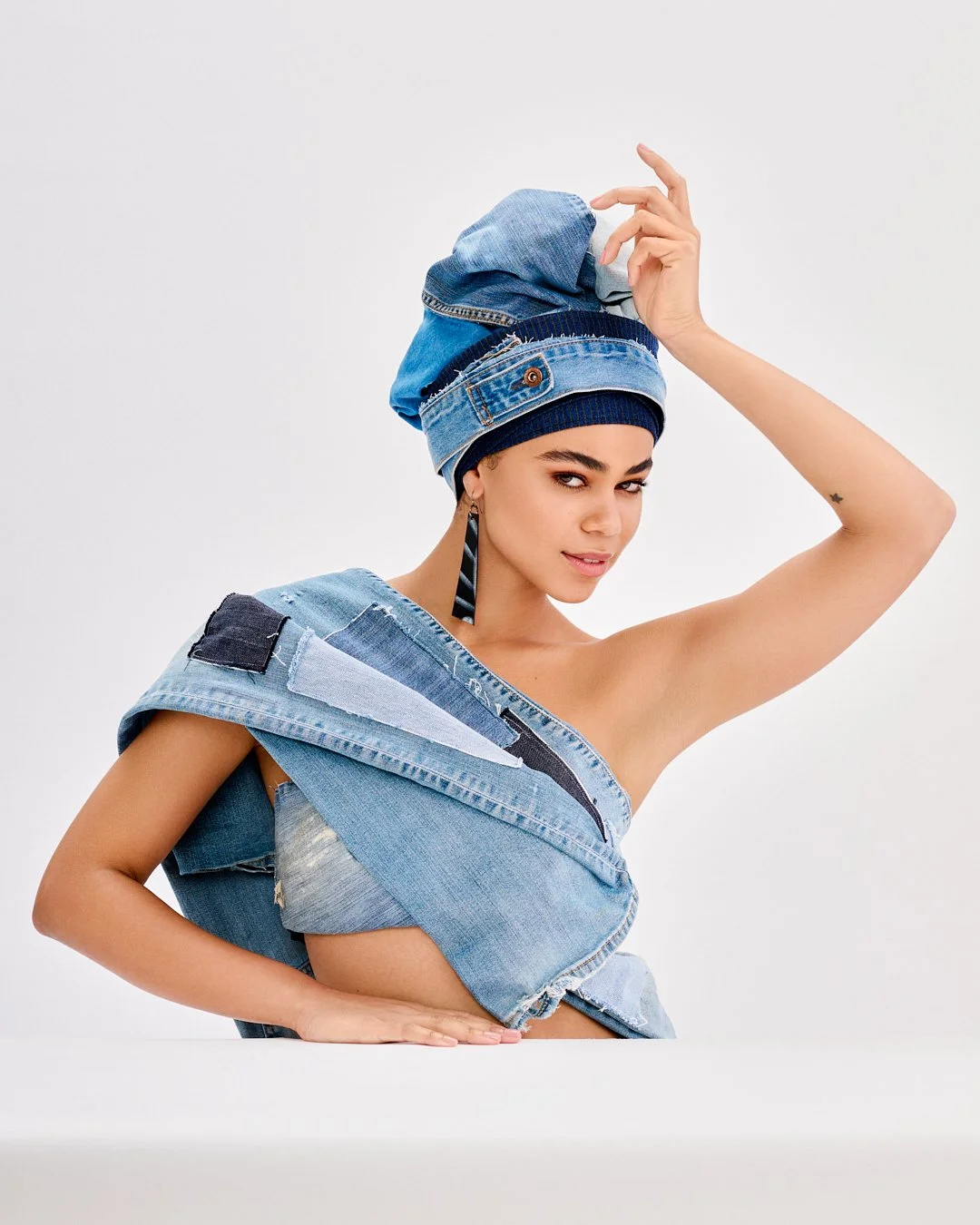 A woman in a creative denim outfit and headwrap, with one arm raised and a confident expression.