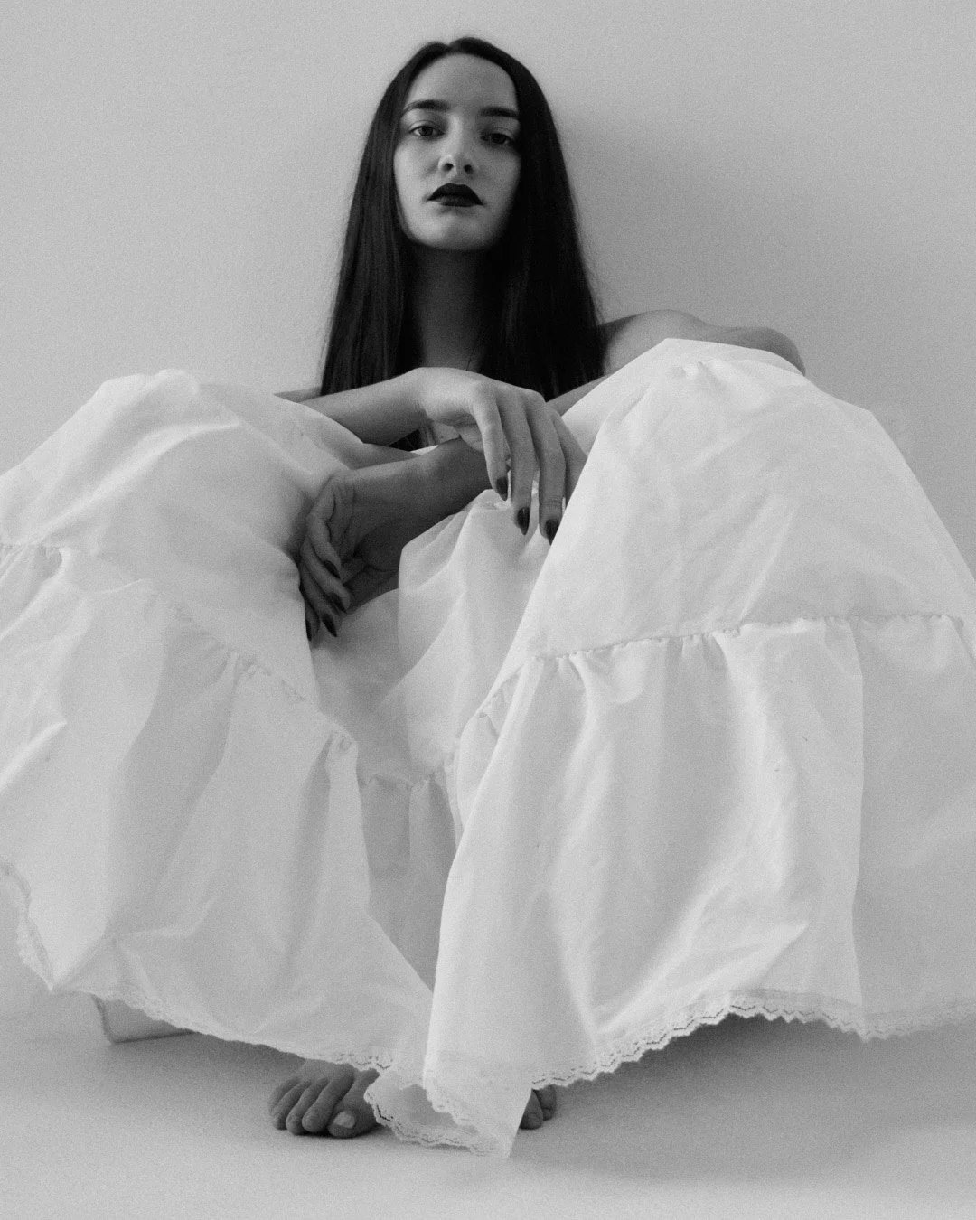Black and white photo of a woman with long dark hair, makeup with dark lipstick, sitting behind a large, crumpled white fabric or paper, with her foot visible at the bottom of the image.