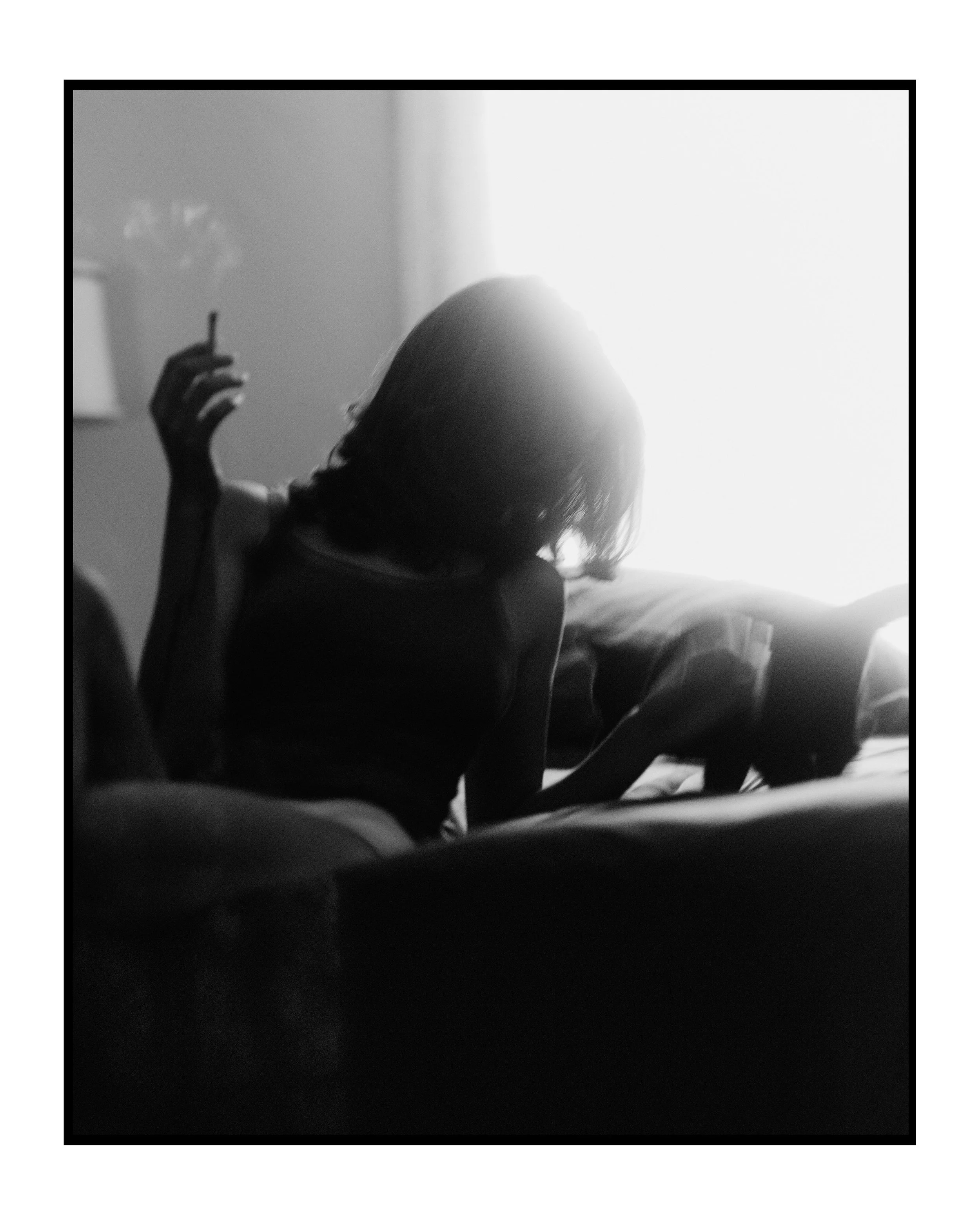 Silhouette of a woman sitting on a bed, smoking a cigarette, with a bright window behind her illuminating her hair and creating a contrast with the dark room.