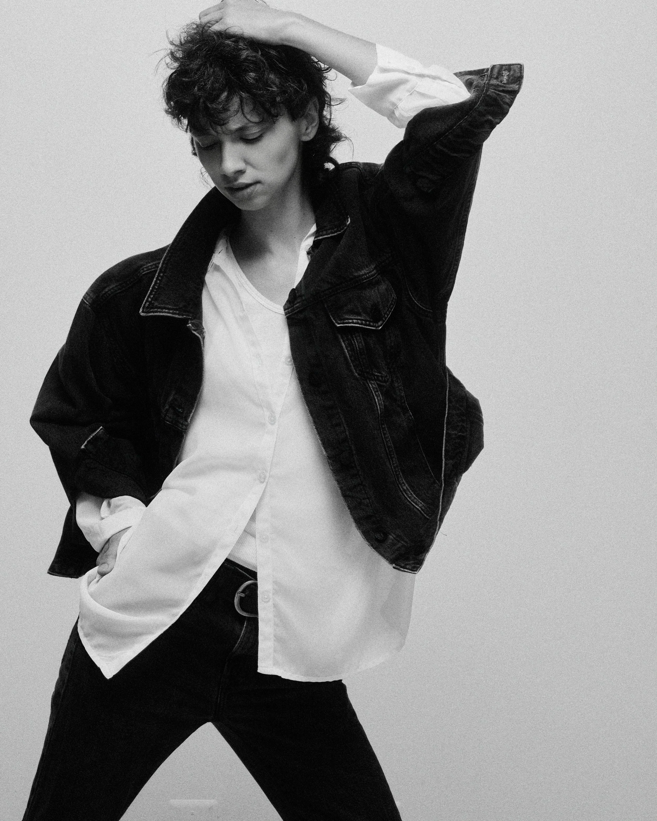 A young woman with curly hair, wearing a white shirt and a denim jacket, posing with her hand on her head against a plain background.