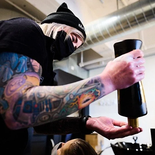 Person with tattoos wearing a black face mask and beanie, holding a black cylindrical object in one hand, indoors with industrial ceiling vents.
