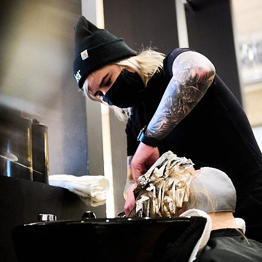 Woman wearing a black beanie and face mask, with visible tattoos on her arm, washing or rinsing hair at a salon sink.