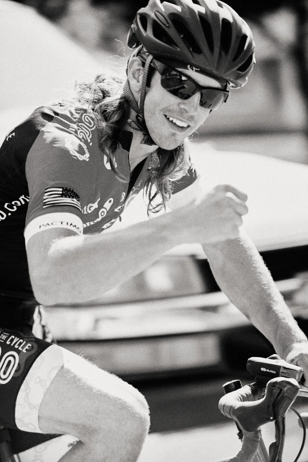 Black and white photo of a smiling cyclist wearing a helmet, sunglasses, and a jersey with a US flag on the sleeve, riding a bike.