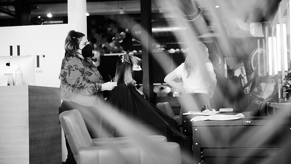 A woman getting a haircut inside a salon, with a stylist wearing a face mask and a woman with long hair seated in a chair, also wearing a face mask, while another stylist works in the background.
