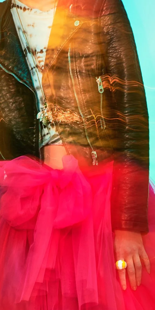 Partially visible person wearing a black leather jacket, pink tulle skirt with a bow, and colorful jewelry, with light streak effects.
