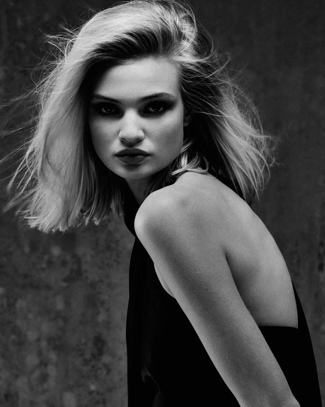 A black-and-white photo of a woman with shoulder-length blonde hair, looking directly at the camera with an intense expression. She is wearing a sleeveless black dress and positioned against a dark, textured background.
