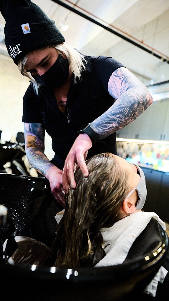 A hairstylist washing a woman's hair in a salon, both wearing face masks.