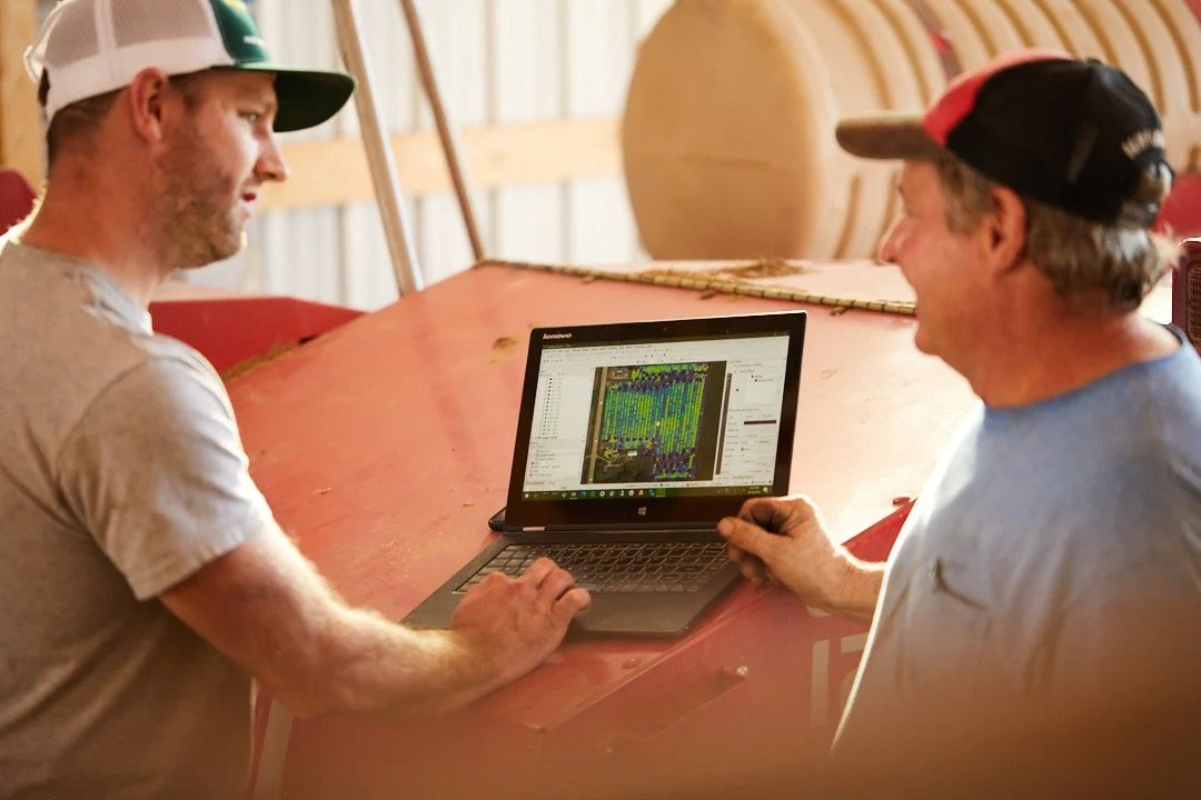 Two men sitting at a wooden workbench in a barn, looking at a laptop with plant growth data or map on the screen.