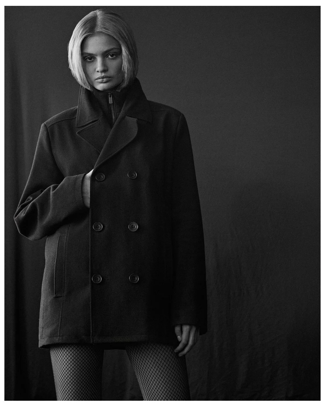 Black and white photo of a woman with short blonde hair wearing a long double-breasted coat and fishnet stockings, standing against a dark background.