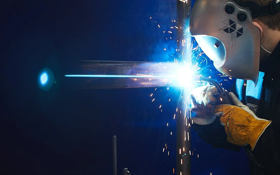 A person welding metal, wearing a protective helmet and gloves, with bright sparks and blue light during welding process.