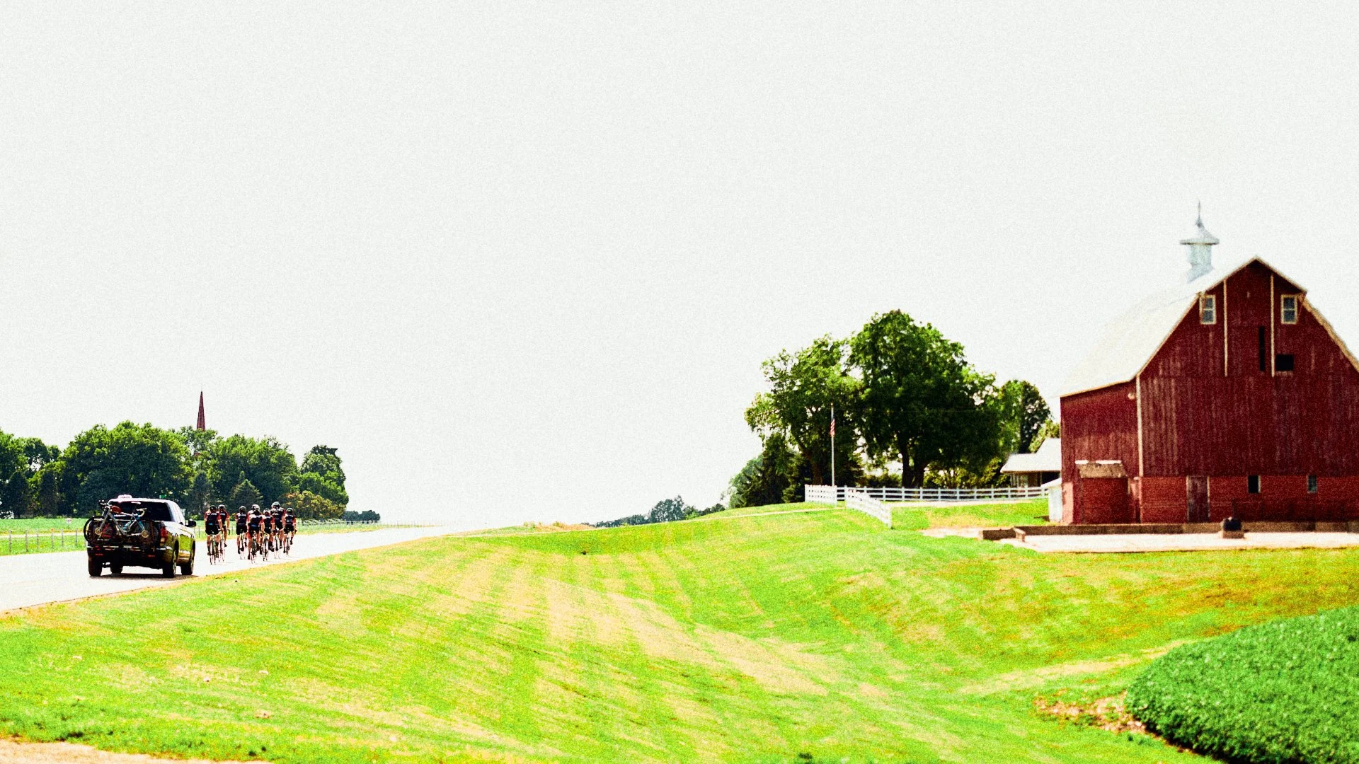 Farmhouse with red exterior, green trees, and white fences on a bright day. Bicyclists and a car on a rural road.