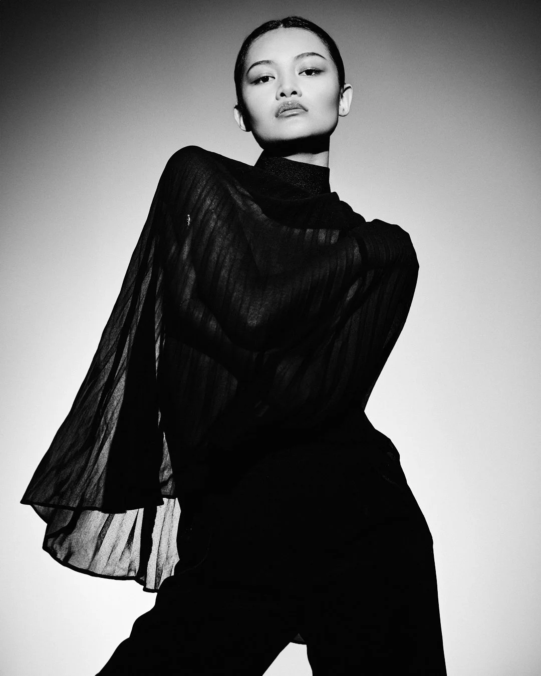 Black and white photo of a woman with short hair wearing a sheer, long-sleeve top and high-waisted pants, posing with one arm crossed over her chest and looking at the camera.