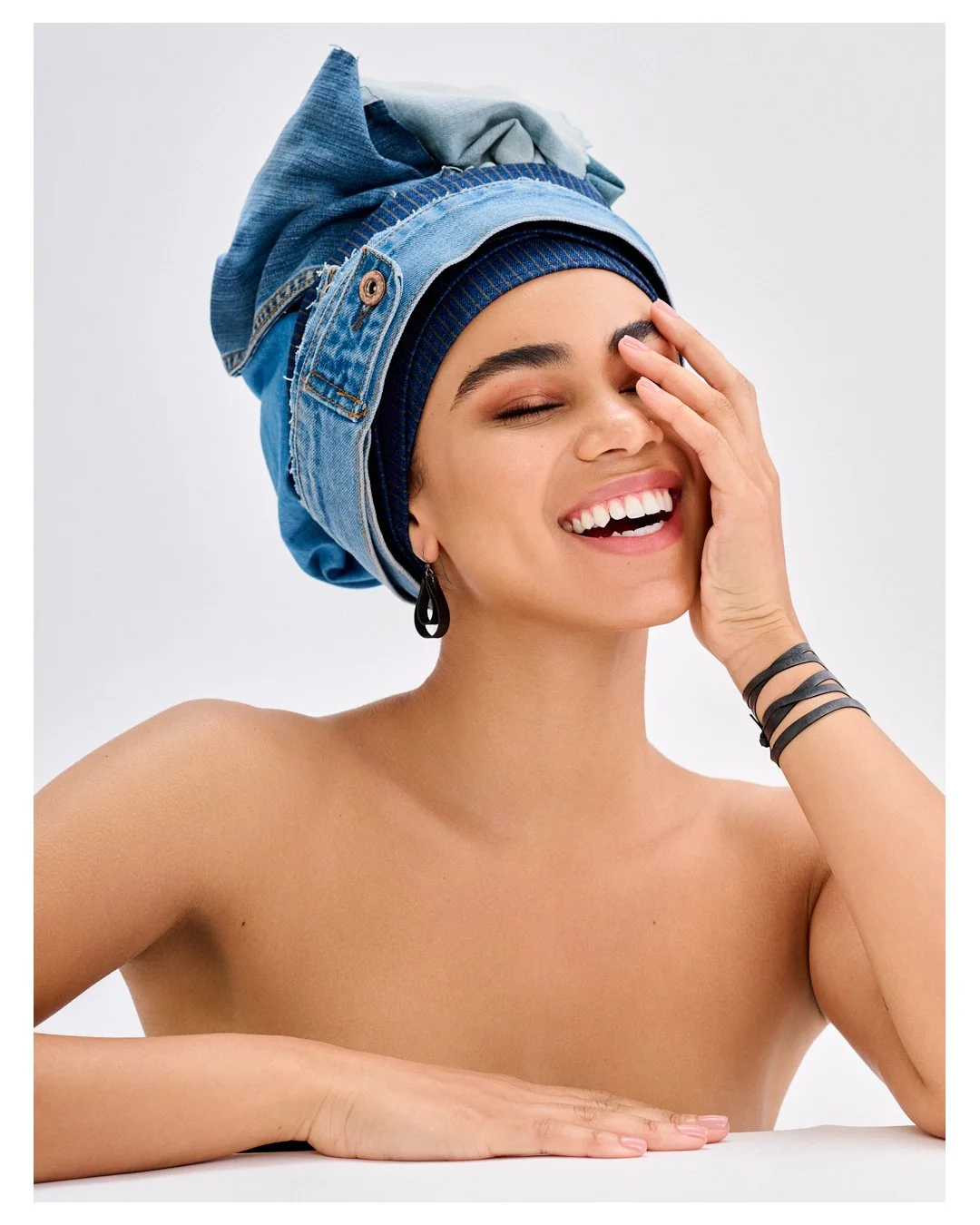 A young woman with a bright smile, wearing multiple black bracelets and a black earring, with her hand covering part of her face. She is topless and has a layered blue headscarf made of denim and fabric, styled in an artistic way, against a plain whi
