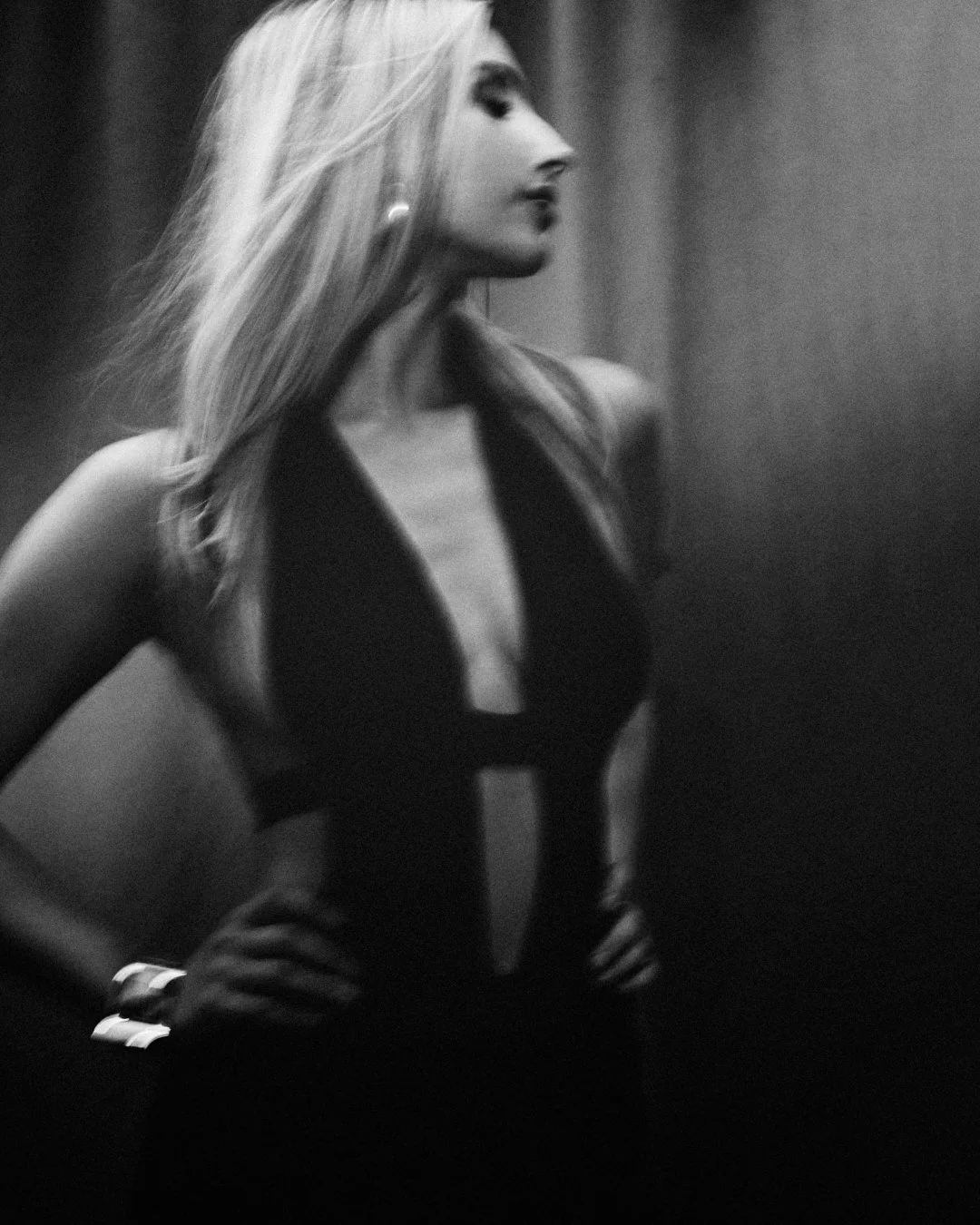 Black and white photo of a woman with long blonde hair, wearing a sleeveless dress with a plunging neckline, standing with hands on hips.
