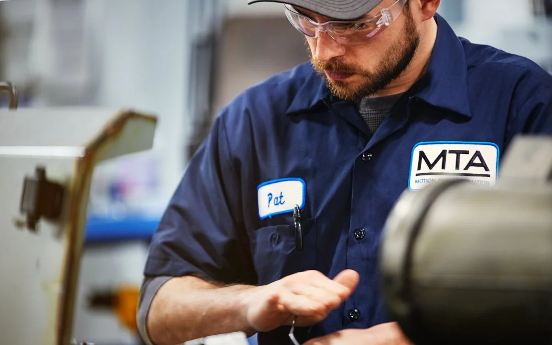A man wearing safety glasses and a navy blue work uniform with the MTA logo and a name tag reading 'Pat' working on machinery.