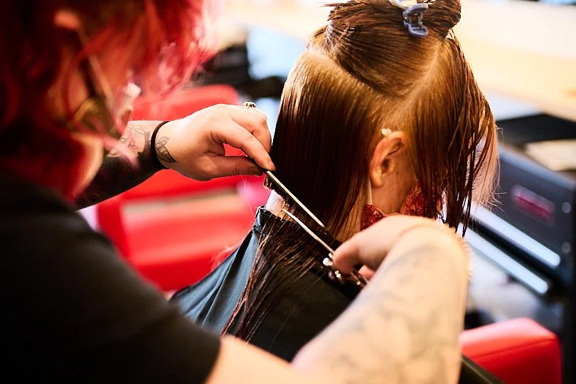 Hairdresser cutting a client's wet hair in a salon with red chairs.