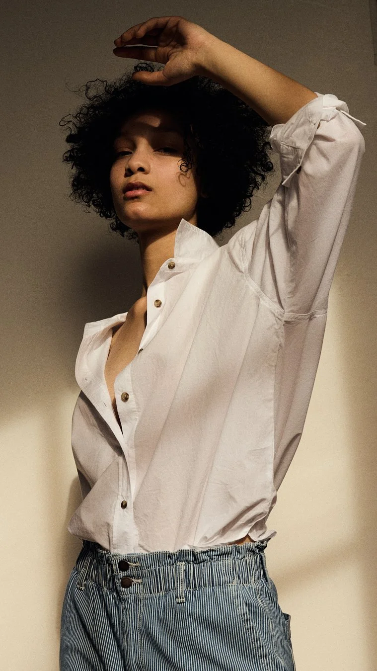 Young woman with curly hair wearing a loose white shirt and striped pants, standing indoors with arm raised.