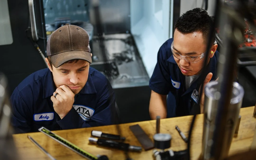 Two young men working on a project at a workbench with tools and equipment, wearing navy blue work uniforms.