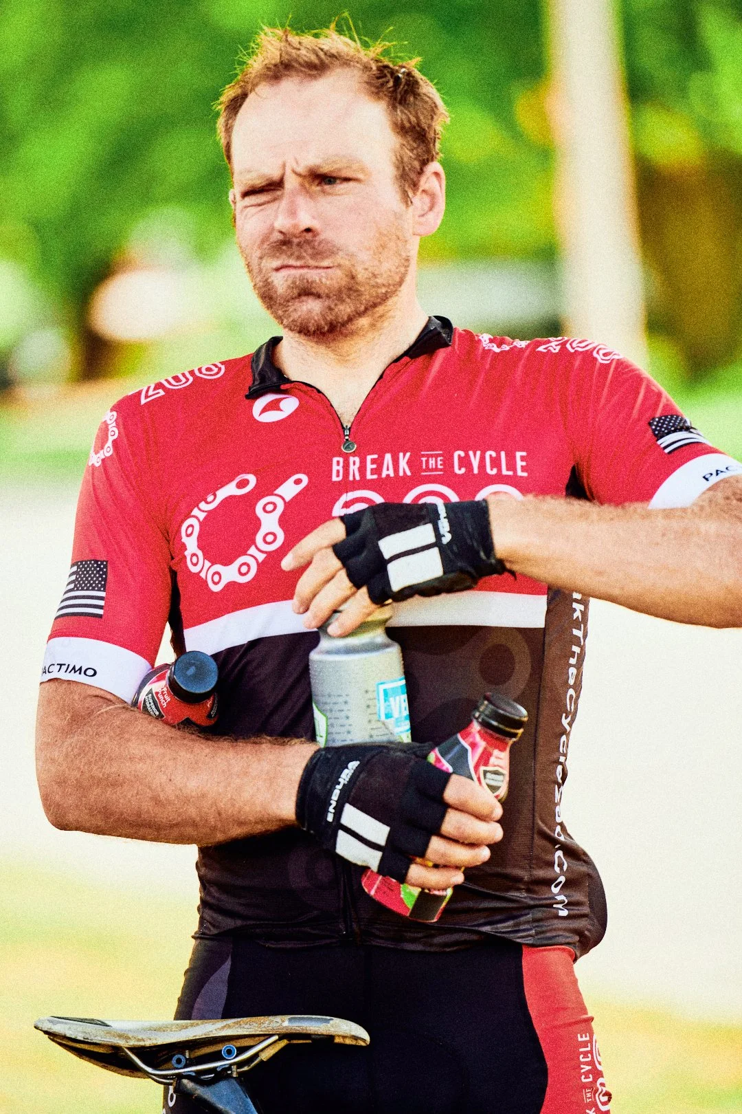 A man wearing a red cycling jersey and gloves, holding water bottles, with a serious expression, standing outdoors with a green background.