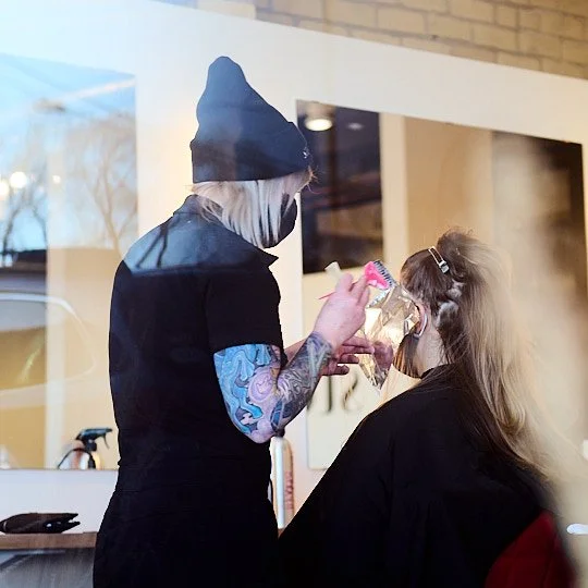 A hairstylist with tattoos, wearing a beanie and mask, working on a woman's hair in a salon.