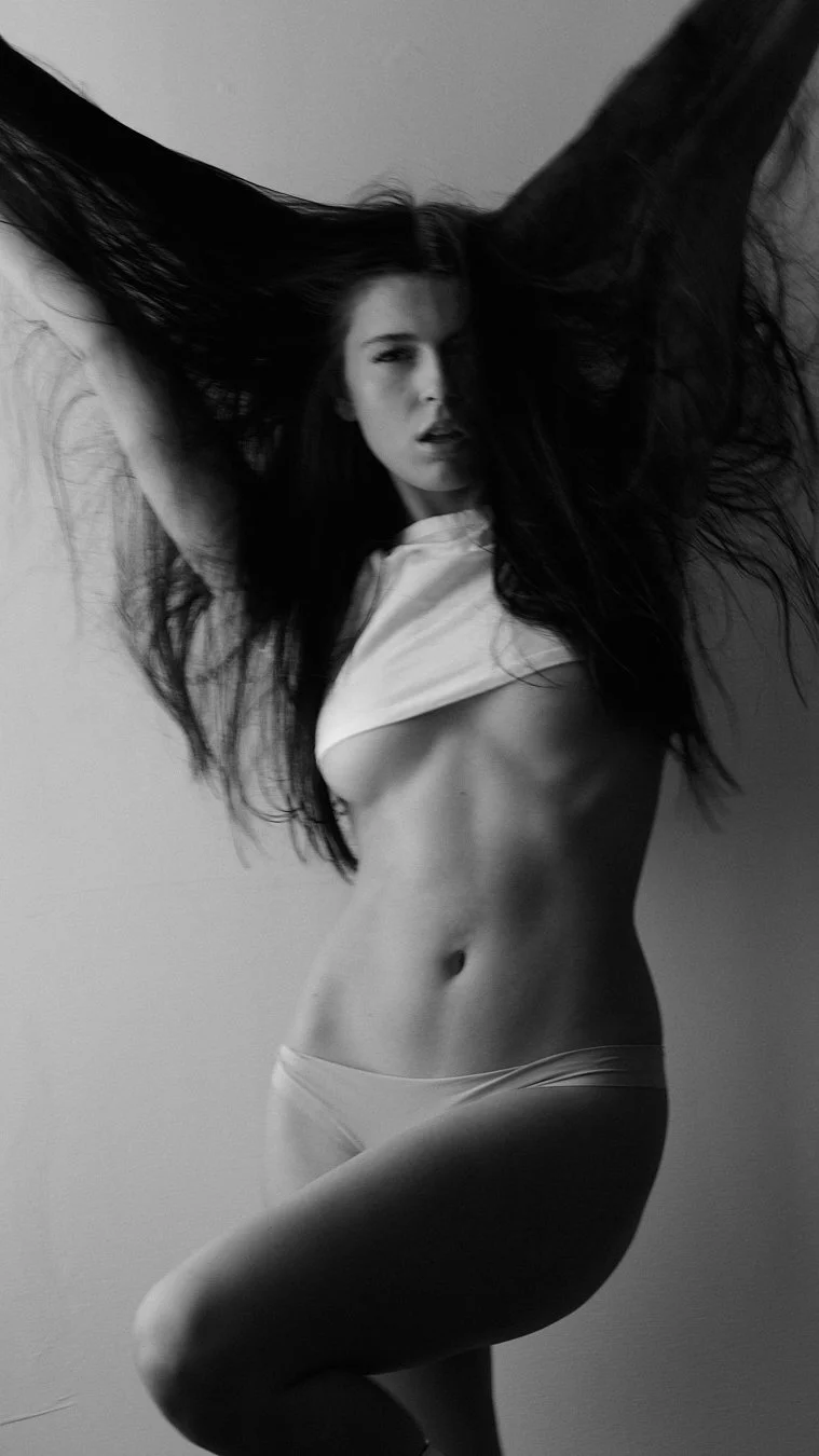 A woman with long dark hair is lifting her arms and wearing minimal clothing, exposing her toned midsection and legs, in a black and white photo.