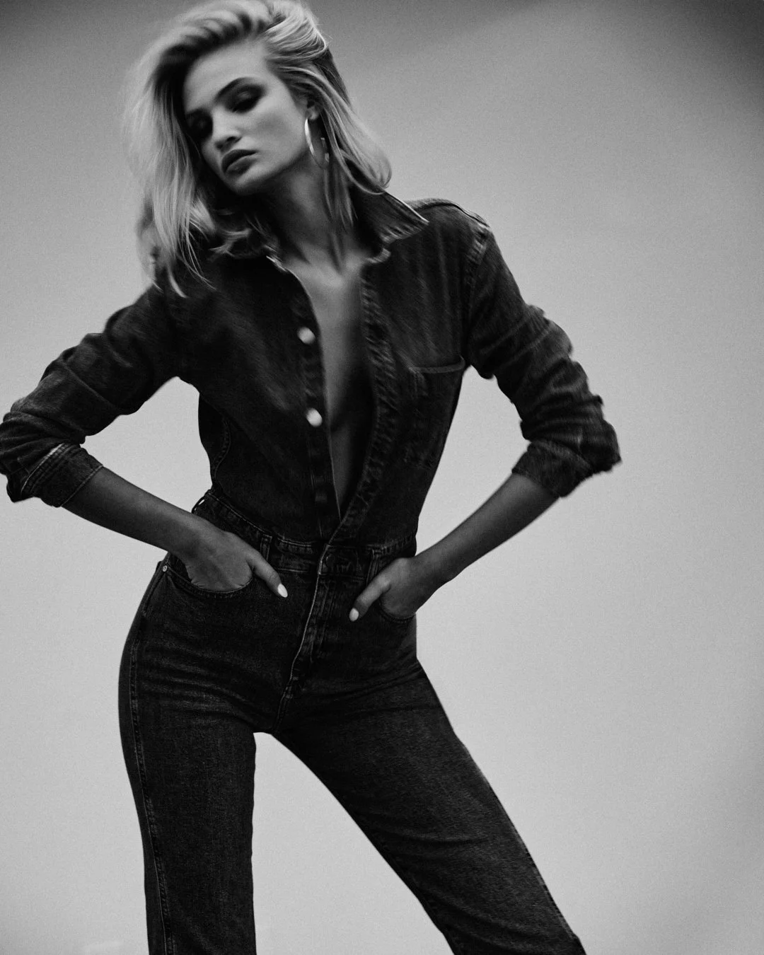 Black and white photo of a woman with blonde hair wearing a denim jacket and jeans, posing with hands in her pockets and leaning slightly to one side.