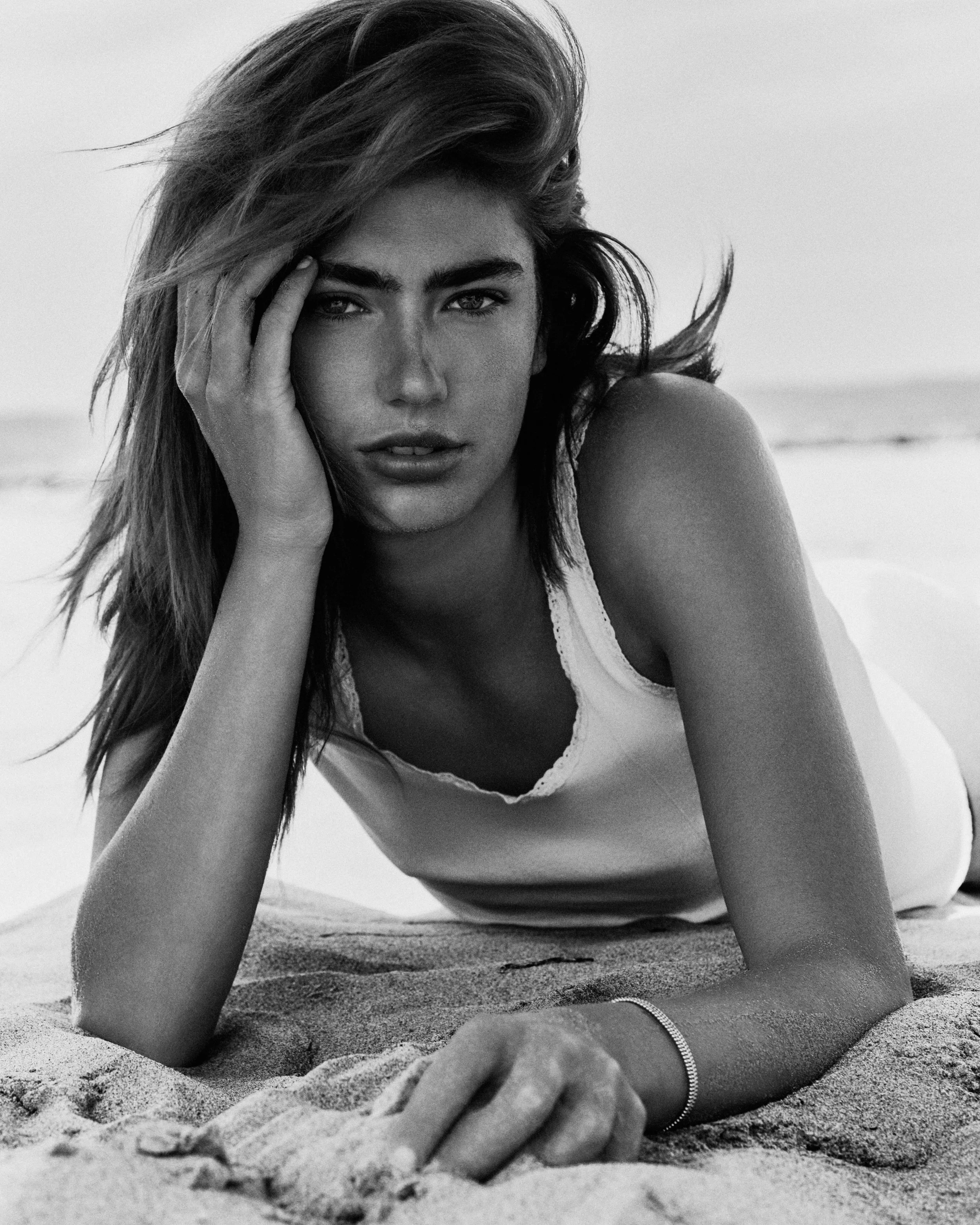 Black and white photograph of a woman lying on the sand at the beach, with one hand resting on her head and the other on the sand, looking directly at the camera.
