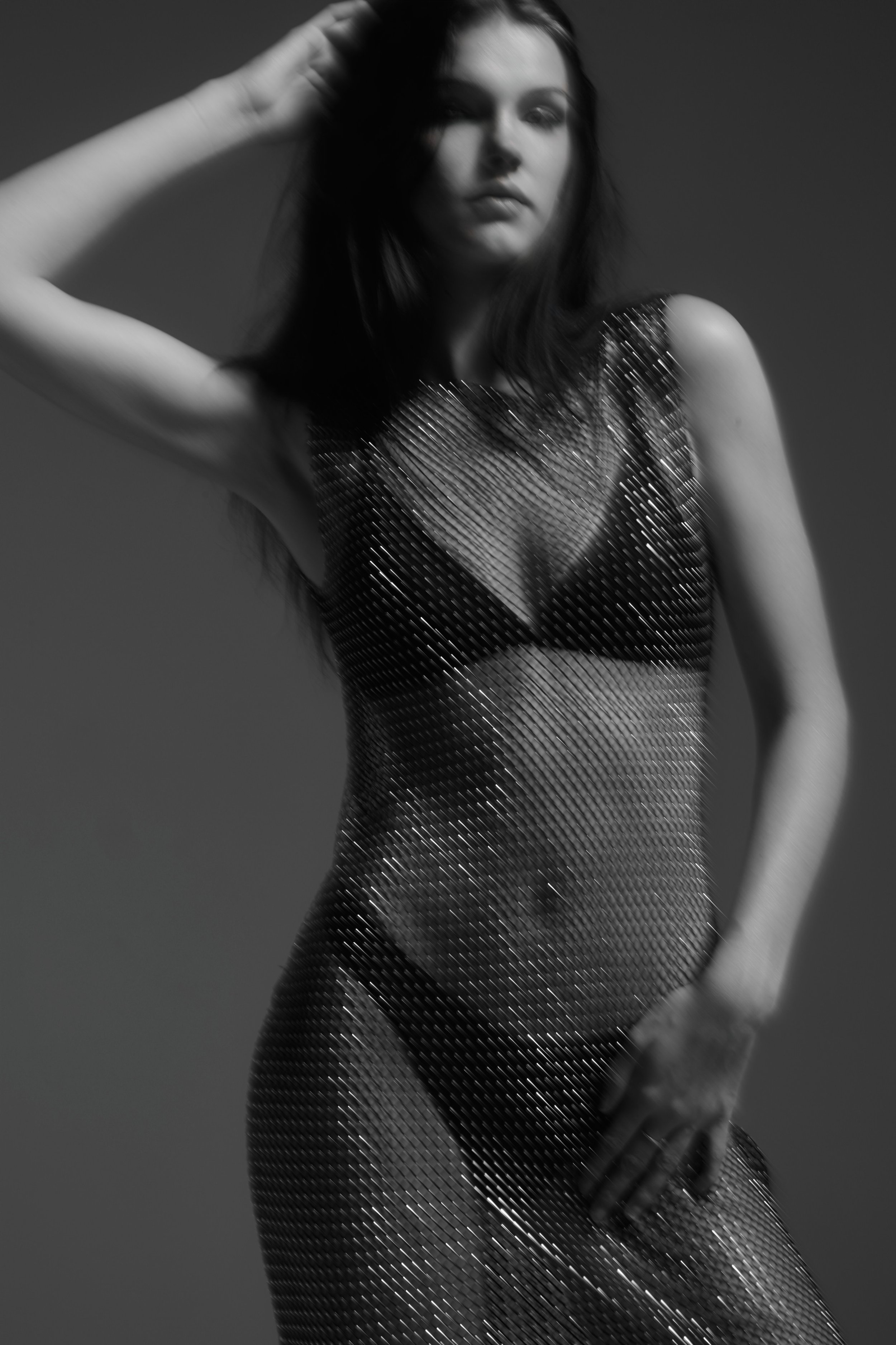 A woman with long dark hair wearing a metallic, sheer dress poses with one hand on her head and the other on her hip, photographed in black and white.