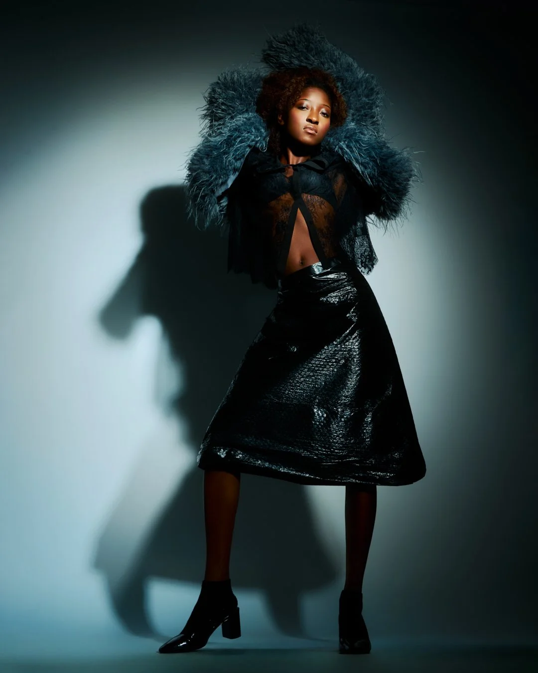A woman posing in a black, shiny skirt, black heeled boots, and a sheer black top, with a large feathered coat over her shoulders, standing against a gray background with shadow and dramatic lighting.